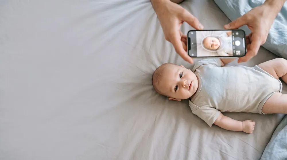 Ein Baby liegt auf einem hellgrauen Laken und wird von oben mit dem Smartphone fotografiert